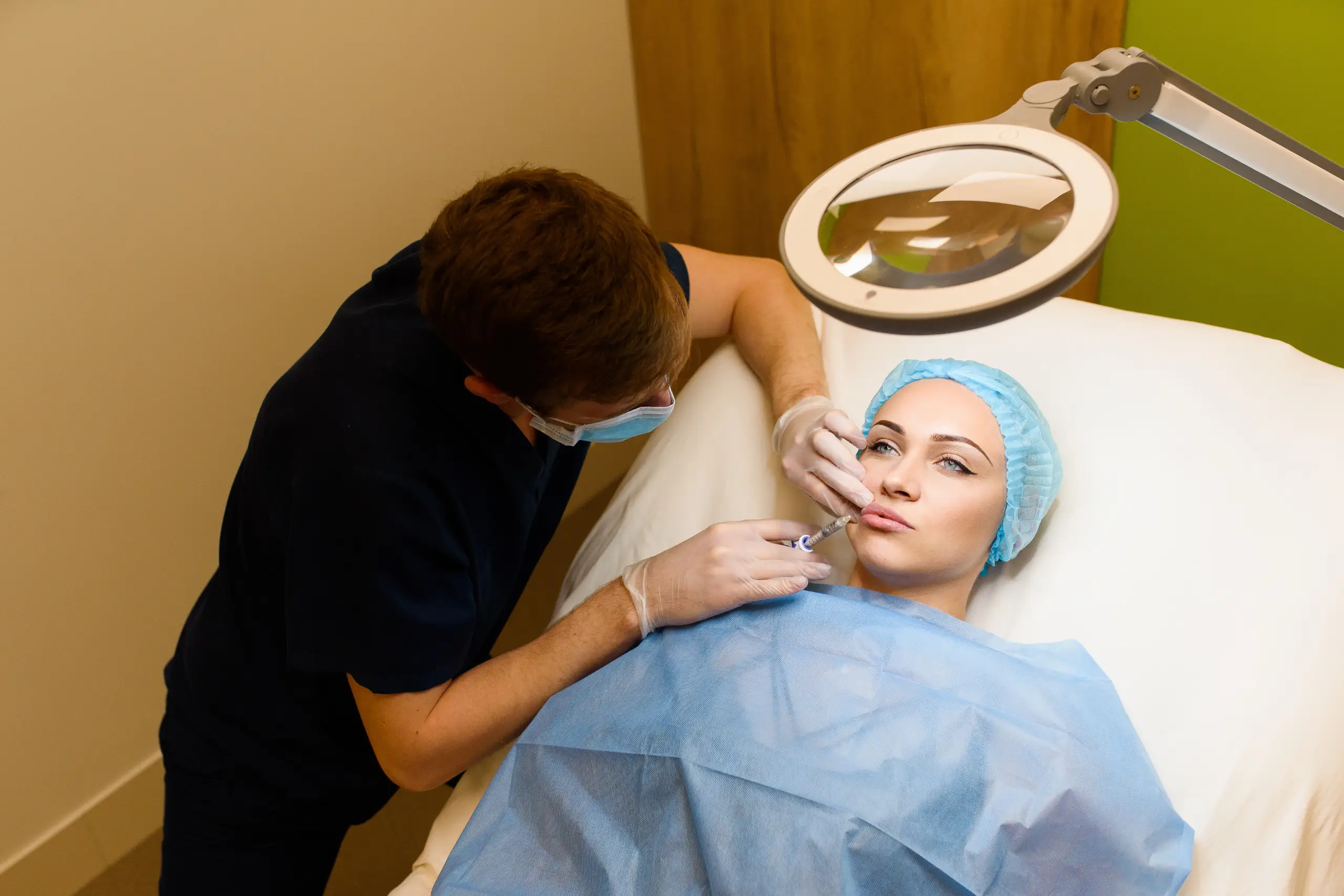 Overhead view of a patient lying in a treatment chair covered with a blue drape while a provider wearing a face mask performs a precise facial injection.